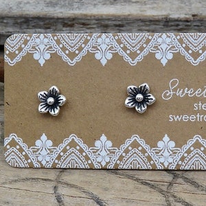 May include: Two small silver flower stud earrings attached to a rectangular brown display card with white lace-style borders. The card features the text Sweet Rock Candy, sterling silver .925, and sweetrockcandy.etsy.com printed in white.
