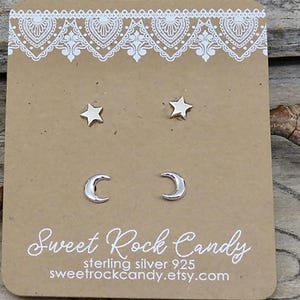 May include: Two pairs of silver stud earrings, one shaped like stars and one shaped like crescent moons, mounted on a rectangular brown display card with white lace-style trim and the text Sweet Rock Candy sterling silver 925.