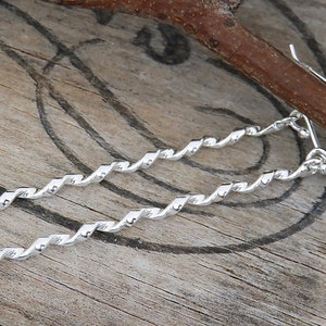 May include: A pair of long, silver-colored earrings featuring a twisted, spiral metal design. Each earring hangs from a simple curved hook, showcasing a reflective, polished surface that catches the light along the length of the spiraled metal bar.