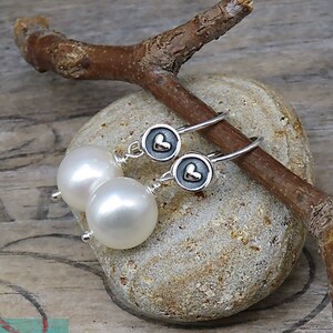 May include: A pair of drop earrings featuring round white pearls suspended from silver hooks. Each earring includes a small circular silver charm with a raised heart detail in the center, connected to the pearl by a thin silver wire wrap.