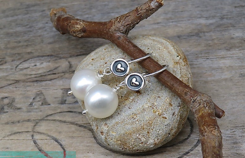 May include: A pair of drop earrings featuring round white pearls suspended from silver hooks. Each earring includes a small circular silver charm with a raised heart detail in the center, connected to the pearl by a thin silver wire wrap.