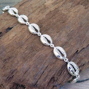 May include: A silver-toned bracelet featuring a series of connected cowrie shell-shaped links. Each link has a polished, reflective surface with a central textured slit, creating a uniform chain design that is linked together in a linear fashion.