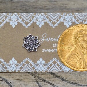 May include: Two silver-colored flower stud earrings on a brown card with white lace-like trim. A penny is next to the earrings. The card has the text "Sweet" and "sweetre".