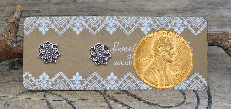 May include: Two silver-colored flower stud earrings on a brown card with white lace-like trim. A penny is next to the earrings. The card has the text "Sweet" and "sweetre".
