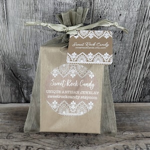 May include: A small gift wrapped in tan paper and a sheer, light gray ribbon. The package has a tag and a sticker with the words "Sweet Rock Candy" and "Unique Artisan Jewelry". The background is a weathered wooden surface.