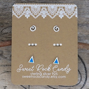 May include: A set of three pairs of stud earrings on a brown display card. The top pair features silver spirals, the middle pair consists of three small silver beads, and the bottom pair displays triangular blue opal stones. Text reads Sweet Rock Candy.