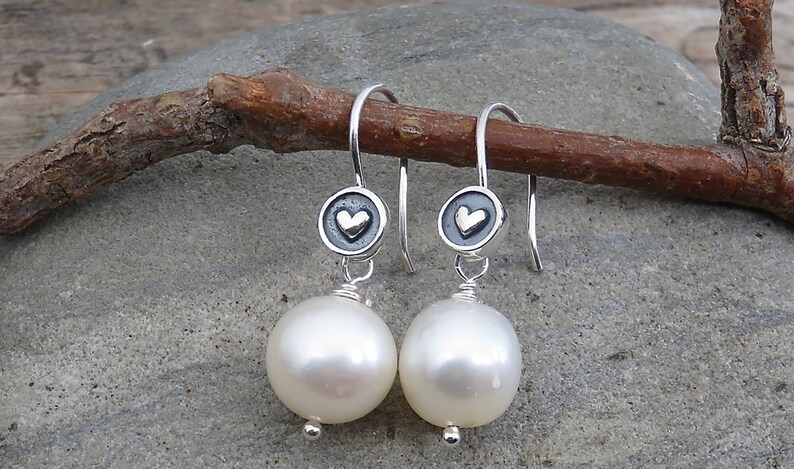 May include: A pair of silver drop earrings featuring a round, dark oxidized silver charm with a small raised heart in the center. Each earring is finished with a large, smooth, white spherical pearl dangling from the bottom of the silver charm.
