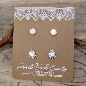 May include: Two pairs of stud earrings on a brown display card. The top pair features simple round white stones. The bottom pair features round white stones set in decorative silver frames. The card displays the text Sweet Rock Candy sterling silver 925.