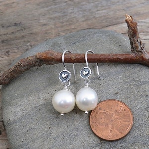 May include: A pair of silver drop earrings featuring round white pearls suspended from small circular charms with a raised heart design. The earrings are displayed next to a copper penny for scale, showing the size of the pearl components.