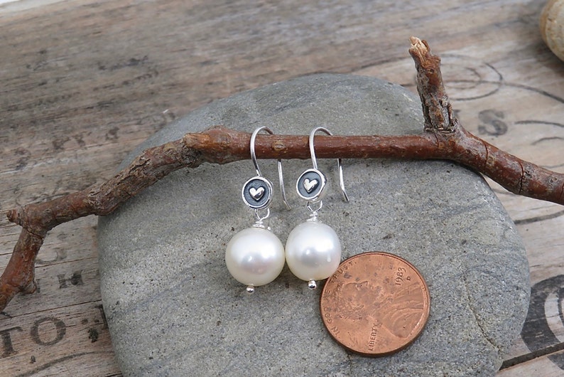 May include: A pair of silver drop earrings featuring round white pearls suspended from small circular charms with a raised heart design. The earrings are displayed next to a copper penny for scale, showing the size of the pearl components.