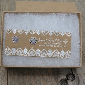 May include: A pair of silver-colored flower stud earrings displayed in a small, open cardboard box filled with white packing material. The earrings are on a brown card with white lace-like trim and the text "Sweet Rock Candy".