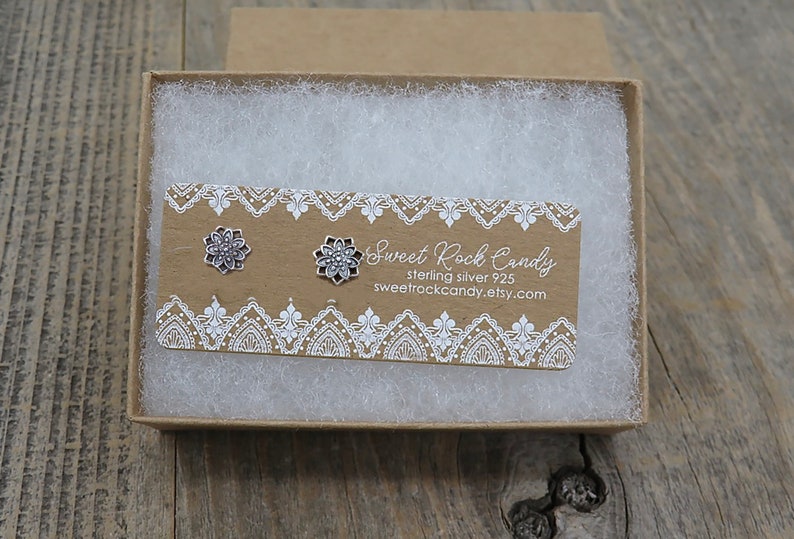 May include: A pair of silver-colored flower stud earrings displayed in a small, open cardboard box filled with white packing material. The earrings are on a brown card with white lace-like trim and the text "Sweet Rock Candy".