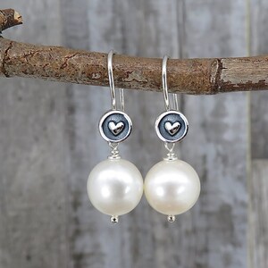 May include: A pair of drop earrings featuring large, round, white pearls suspended from silver hooks. Each earring includes a small, circular, oxidized silver charm with a raised, polished silver heart in the center, connecting the hook to the pearl.