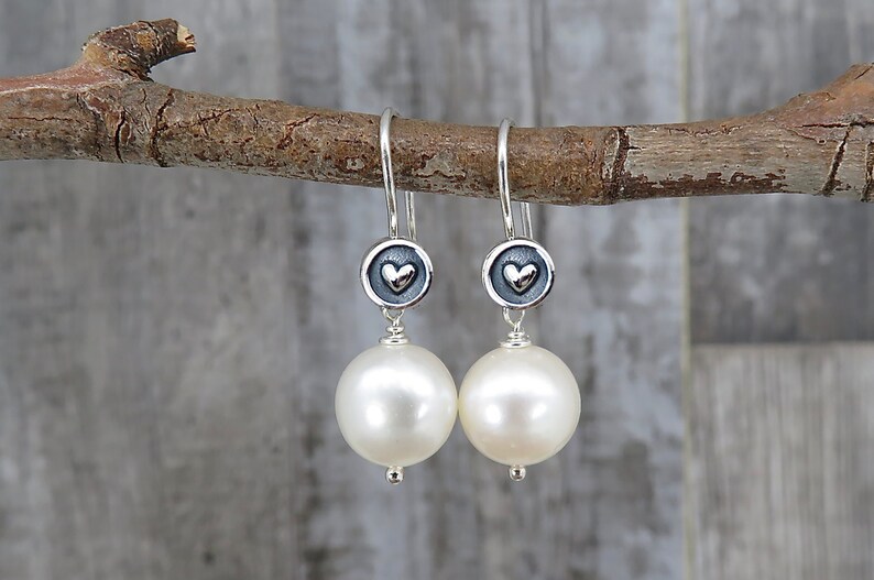 May include: A pair of drop earrings featuring large, round, white pearls suspended from silver hooks. Each earring includes a small, circular, oxidized silver charm with a raised, polished silver heart in the center, connecting the hook to the pearl.
