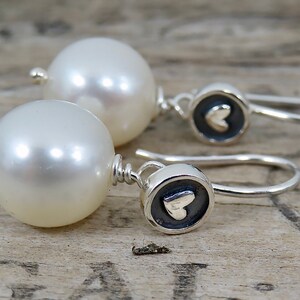 May include: A pair of drop earrings featuring large, round, white pearls suspended from silver hooks. Each earring includes a small, circular silver charm with a dark, recessed background and a raised, polished silver heart in the center.