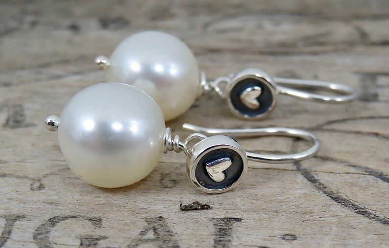 May include: A pair of drop earrings featuring large, round, white pearls suspended from silver hooks. Each earring includes a small, circular silver charm with a dark, recessed background and a raised, polished silver heart in the center.