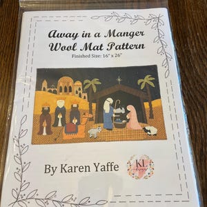 May include: A packaged wool mat pattern titled "Away in a Manger." The finished size is 16" x 26". The pattern features a colorful illustration of the nativity scene with the text "By Karen Yaffe" and a logo.