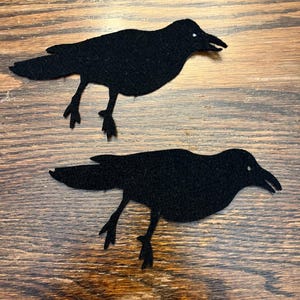 May include: Two black felt crow cutouts with white eye details. The crows are in a walking pose, with their legs and beaks visible. The background is a wooden surface.