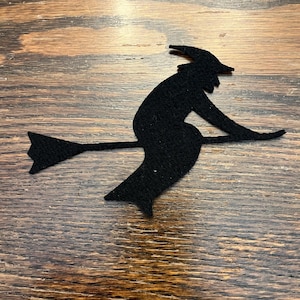 May include: A black felt silhouette of a witch riding a broomstick. The witch is wearing a pointed hat and a long dress. The broomstick extends horizontally, and the witch is angled slightly forward, suggesting movement. The background is a wooden surface.