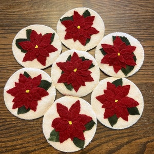 May include: Seven white felt coasters with red poinsettia flower designs and green leaves. The coasters are arranged in a circle on a wooden surface.