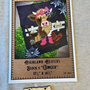 May include: A framed fabric art piece titled "Highland Heifers Block 5 'Ginger'" featuring a cow wearing a pink bow and boots, with a chicken and a fence. The artwork measures 12.5 x 14.5 inches. A wooden sign that says "Ginger" is below the artwork.