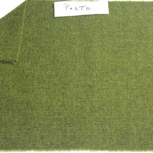 May include: A rectangular piece of olive green fabric with a textured, woven appearance. The top left corner is folded over to reveal the underside. A white rectangular label is attached to the top center with the handwritten text Pesto.