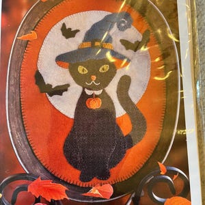 May include: A craft pattern titled Practical Magic by Sew Cherished featuring a black cat wearing a blue witch hat and a small orange pumpkin charm around its neck, set against a white moon and orange background inside an oval frame.