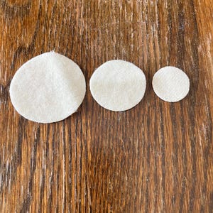 May include: Three white felt circles of different sizes on a brown wooden surface.