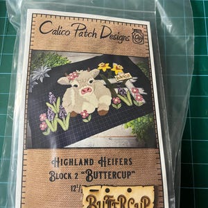 May include: A packaged craft kit featuring a Highland Heifers design. The kit includes a printed image of two cows with floral accents, the text "Calico Patch Designs", and a wooden cutout that says "Buttercup". The block size is 12 1/2 inches.