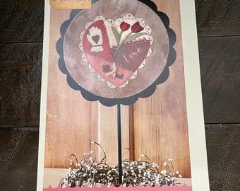 May include: A craft project featuring a heart-shaped design with red, pink, and green elements, including the word "Love." The design is set against a circular backdrop with a scalloped edge and a black stem. The text "A Round the Year February" is visible.