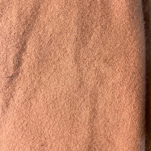 May include: Close-up of a peach-colored fabric with a textured, slightly fuzzy surface. The fabric appears to be a solid color with subtle variations in tone, suggesting a natural fiber. The material is folded, creating shadows and depth.
