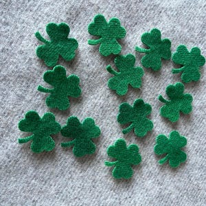 May include: A collection of eleven green felt shamrock shapes, each with three leaves and a stem. The shamrocks are scattered on a gray, textured fabric background, perfect for St. Patrick's Day crafts.