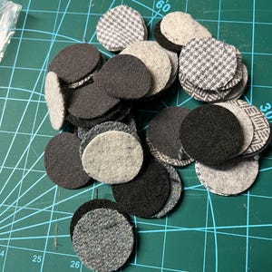 May include: A collection of small, circular fabric pieces in various shades of gray and black. Some pieces have a solid color, while others feature patterned designs like houndstooth or a subtle weave. The pieces are scattered on a green cutting mat.