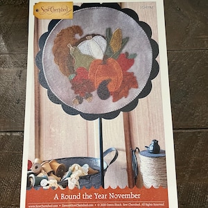 May include: A craft kit titled "A Round the Year November" featuring a felt applique design with a Thanksgiving theme. The design includes a turkey, pumpkins, corn, and fall leaves. The kit is displayed on a wooden background.
