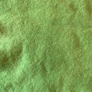 May include: A close-up view of a vibrant lime green felted fabric. The material displays a dense, fuzzy, and soft texture with short, matted fibers creating a uniform, non-directional surface appearance across the entire frame of the image.