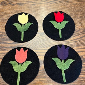 Peut inclure: Quatre dessous-de-verre en feutre avec un fond noir et un motif de tulipe unique en différentes couleurs : jaune, rouge, orange et violet.