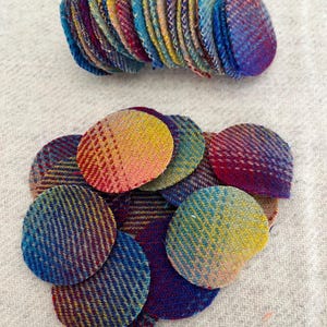 May include: A collection of colorful, round fabric pieces in shades of blue, purple, yellow, and pink. The pieces have a woven texture and are arranged in two groups, one stacked and the other scattered.