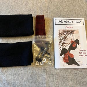 May include: A craft kit containing two rectangular pieces of black fabric, a strip of red plaid fabric, a small clear plastic bag with four white buttons and black hardware, and a white instructional booklet titled All About Ewe Crows.