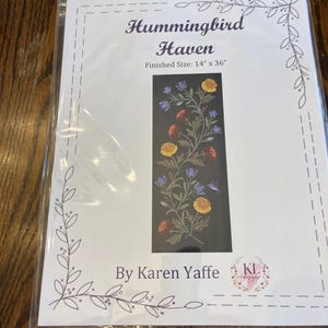 May include: A packaged craft kit titled "Hummingbird Haven" with a finished size of 14" x 36". The design features a floral pattern with red, yellow, and blue flowers on a dark background. The kit is by Karen Yaffe.