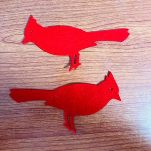 May include: Two red felt cardinal bird cutouts. The birds are facing the right side of the image. The birds are on a woodgrain surface.