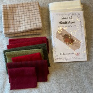 May include: A craft kit titled Star of Bethlehem by Karen Yaffe with a finished size of 12.25 inches by 30 inches, accompanied by a stack of beige plaid fabric and several rectangular swatches of red, dark red, and olive green wool fabric.