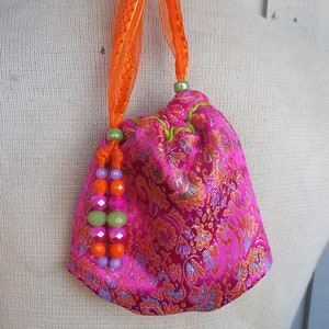 May include: A small pink drawstring bag with a floral pattern. The bag has a green drawstring and an orange ribbon handle. The bag is decorated with pink, green, and orange beads.
