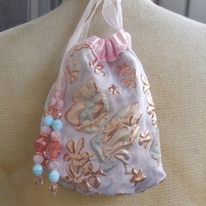 Peut inclure: Un petit sac à cordon rose avec un motif floral doré et un ruban de satin rose. Le sac a un pompon à perles avec des perles roses, bleues et transparentes.