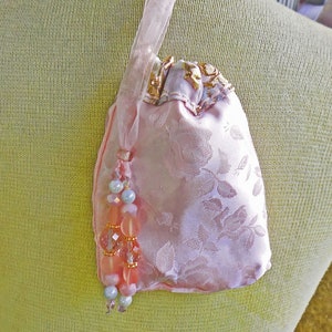 Peut inclure: Un sac à cordon en satin rose avec un motif floral et un pompon perlé. Le sac a une bordure dorée et un ruban blanc.