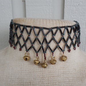 May include: A black and green beaded choker necklace with gold bells hanging from the bottom. The necklace is on a white mannequin.