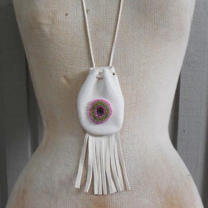 Może przedstawiać: Biały skórzany naszyjnik z sakiewką z okrągłym wzorem z koralików w kolorze różowym, zielonym i fioletowym. Sakiewka ma frędzle na dole i długi biały sznurek. Naszyjnik jest prezentowany na manekinie.