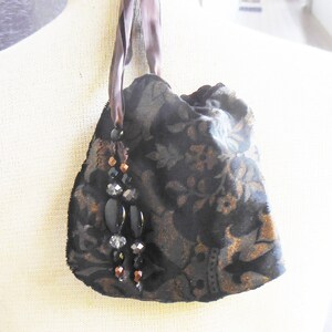 May include: A small black velvet bag with a gold floral pattern and a brown ribbon. The bag has a beaded tassel with black and clear beads.