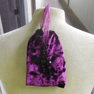 May include: A small purple velvet pouch with a black beaded embellishment. The pouch has a drawstring closure and a ribbon handle.