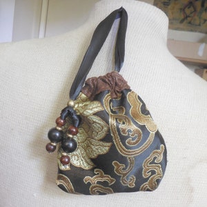 May include: A small black and gold drawstring bag with a brown fabric lining. The bag has a black ribbon handle and is decorated with a floral pattern. The bag has a tassel made of black and brown beads.
