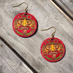 May include: A pair of round wooden earrings with a red background and a carved yellow face design. The earrings have gold-colored hooks and are displayed on a weathered wooden surface. The design features a stylized face with intricate details.
